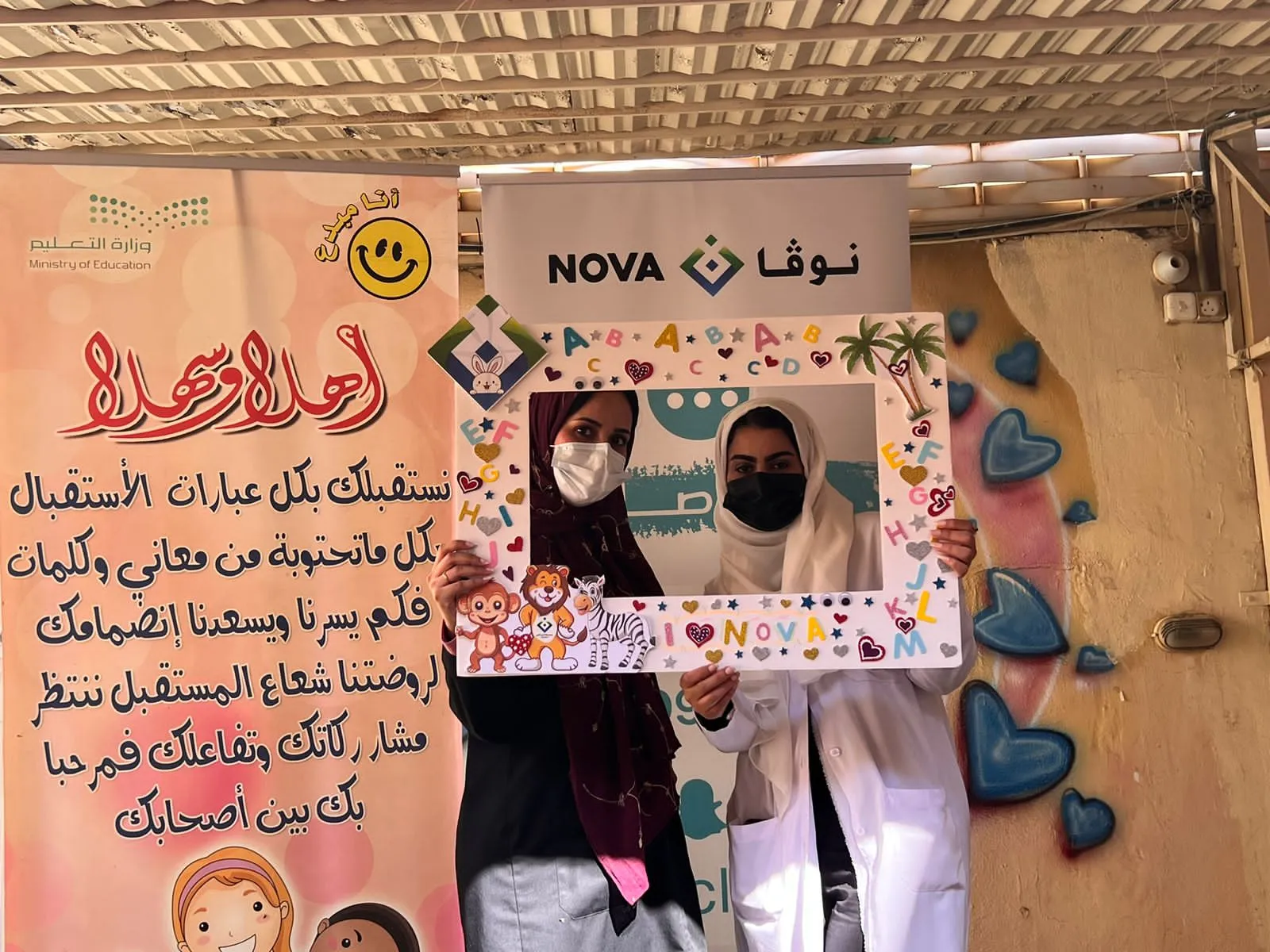 عضوتان من فريق نوفا أمام لافتة الترحيب بالروضة ولافتة المجمع